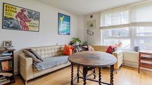 Ein Wohnzimmer mit Sofa, Couchtisch und Bücherregal. An den Wänden hängen Poster und ein Fenster mit Jalousien.