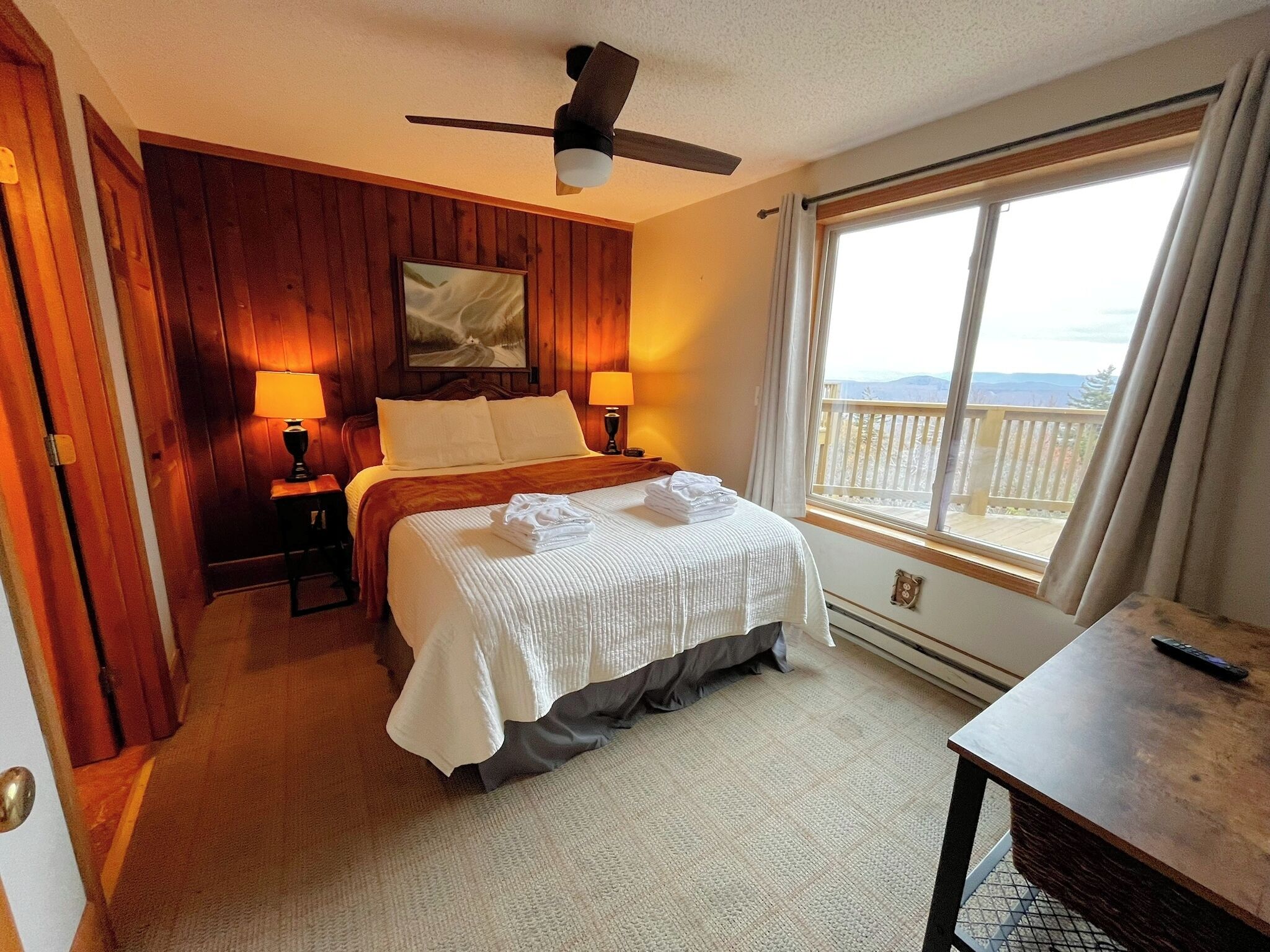 A hotel room with a bed, two bedside lamps, a ceiling fan, a large window with a view, and a wooden door.