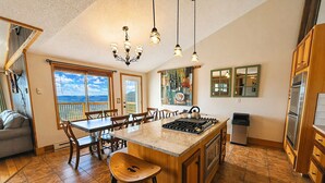 A kitchen with a large island, wooden cabinets, a gas stove, and a dining area with chairs. - Land's End 3 - The Yeti's Den - Snowshoe, Sunset Views! (Snowshoe)