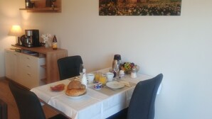 Mesa para café da manhã com cadeiras, máquina de café, tigela de frutas e um quadro na parede.