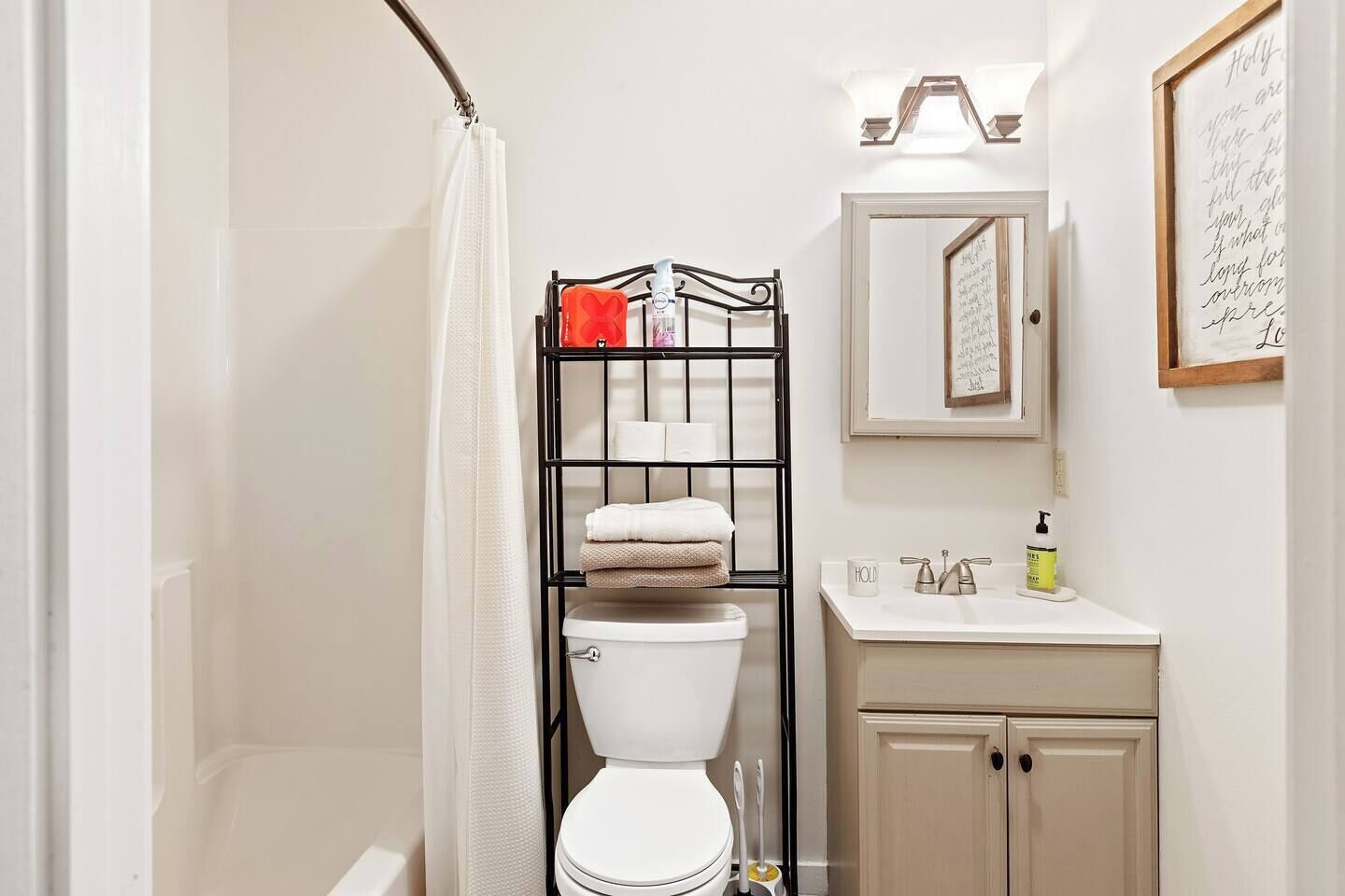 A bathroom with a white toilet, a black metal shelf with towels, a white sink with a mirror above it, and a framed quote on the wall.