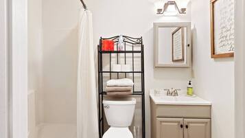 A bathroom with a white toilet, a black metal shelf with towels, a white sink with a mirror above it, and a framed quote on the wall.