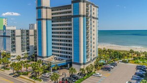 A multi-story hotel with a modern design, located near a beach with a parking lot and palm trees. - SIPPING ON SUNSHINE/OCEANFRONT/PRIME LOCATION (Myrtle Beach)