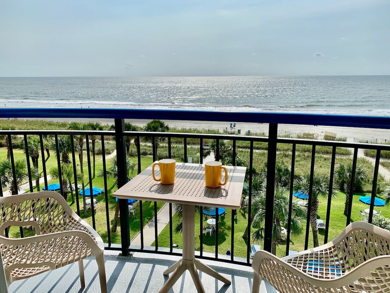 A balcony with a table and two chairs, overlooking a beach and a pool area.