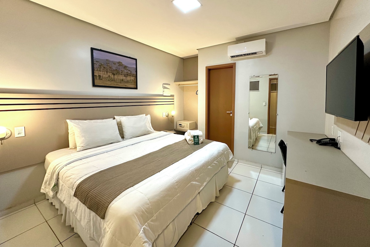 A hotel room with a large bed, a flat-screen TV, an air conditioning unit, and a bathroom mirror.