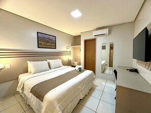 A hotel room with a large bed, a flat-screen TV, an air conditioning unit, and a bathroom mirror.