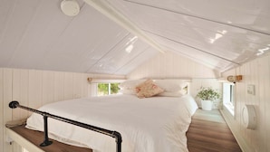 A bedroom with a wooden floor, white walls, a bed with white bedding, a window, and a potted plant.