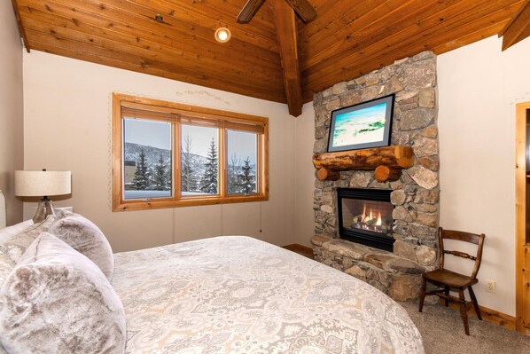 Ein Schlafzimmer mit einem großen Bett, einem Kamin aus Stein, einem Fenster mit Blick auf Schnee und einer Holzbalkendecke.