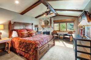 A bedroom with a large bed, a stone fireplace, a wooden ceiling, and a view of the outdoors.