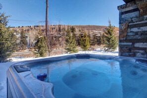 A hot tub with a stone wall on one side, surrounded by snow and trees, with a view of a residential area in the background.