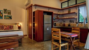 Una camera compatta con un letto, una panca di legno e un tavolino. Accanto, una piccola cucina con un frigorifero, scaffali e una finestra.