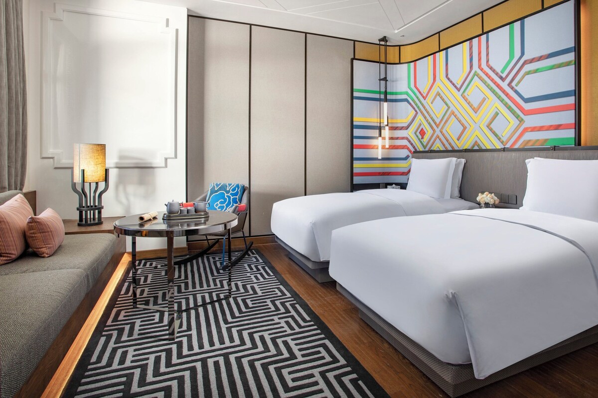 Una habitación de hotel con cama, dos mesitas de noche, un sofá, una alfombra estampada y un mural de colores en la pared.