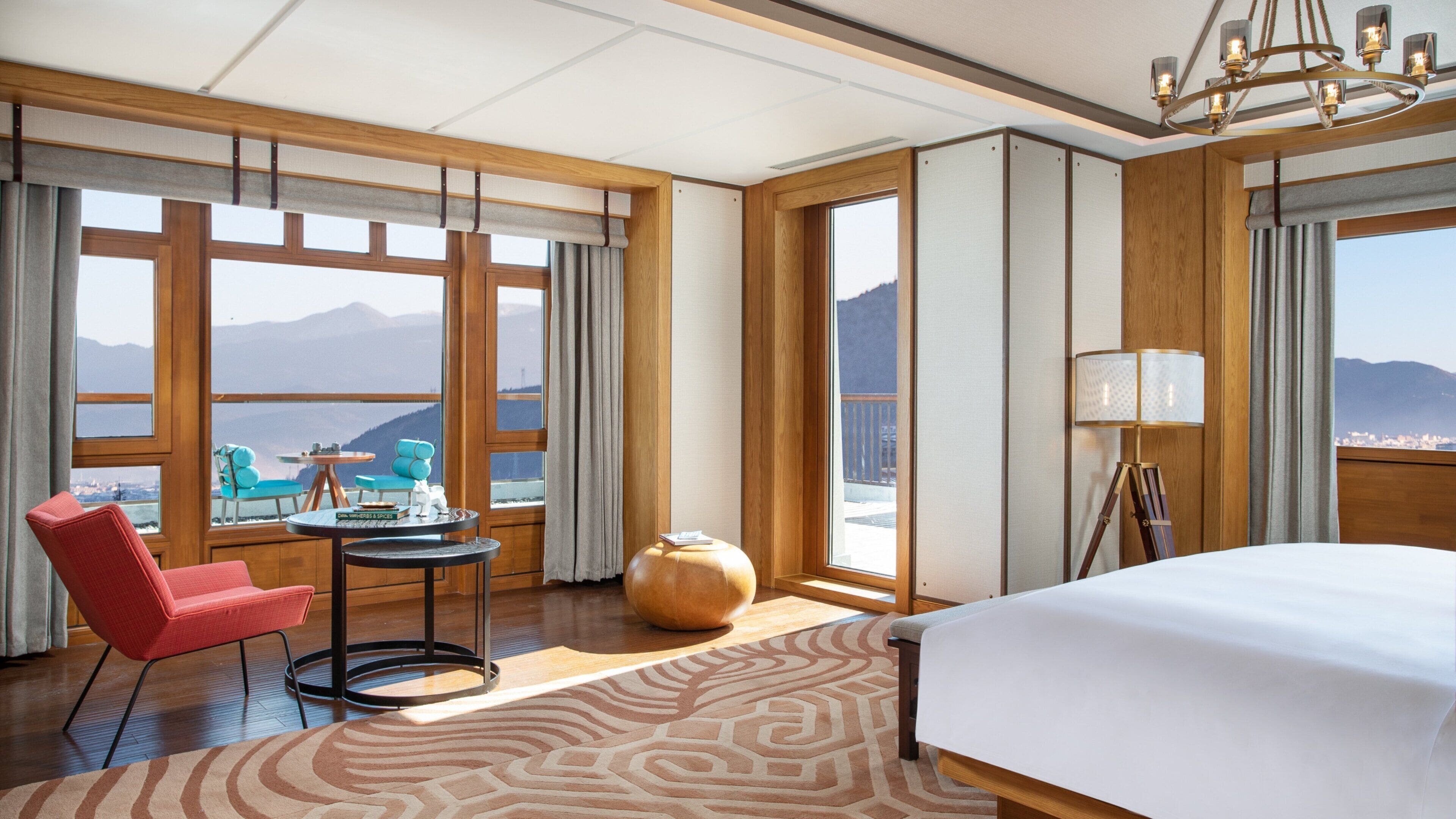 A hotel room with a bed, a desk, a chair, a lamp, and large windows offering a mountain view.