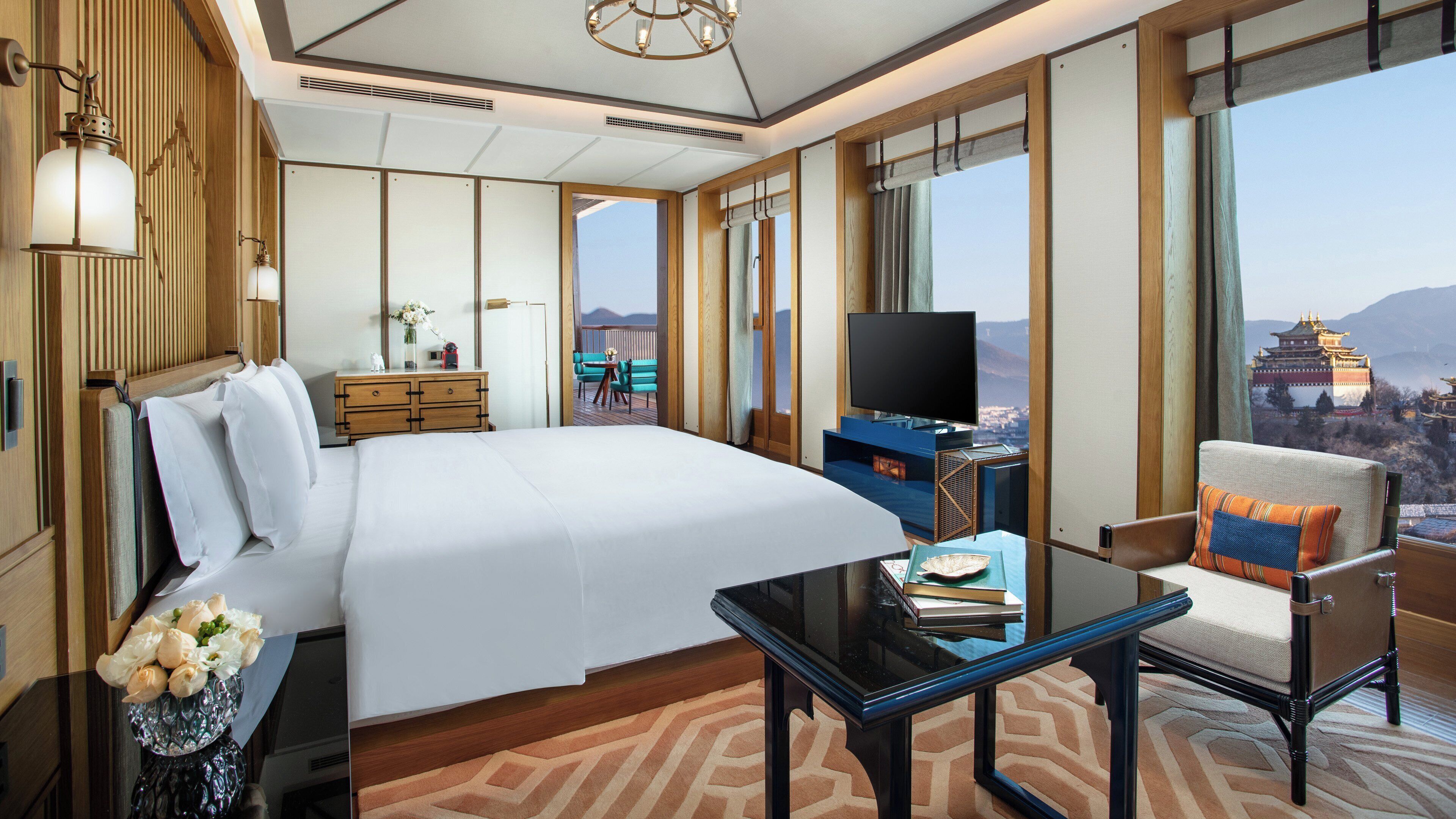 Ein Hotelzimmer mit einem großen Bett, einem Fernseher, einem Schreibtisch und Blick auf eine Berglandschaft sowie traditioneller Architektur.