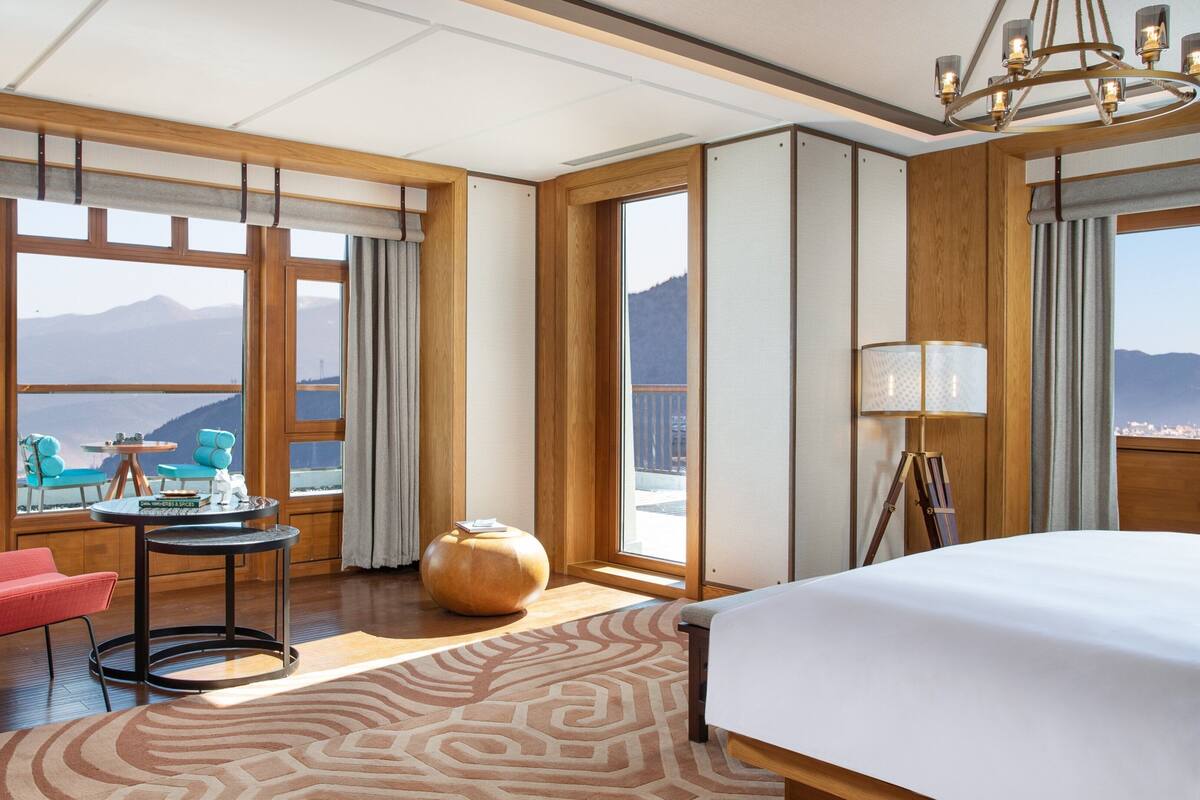 Une chambre d’hôtel équipée d’un lit, d’un bureau, d’une chaise, d’une lampe et de grandes fenêtres donnant sur la montagne.