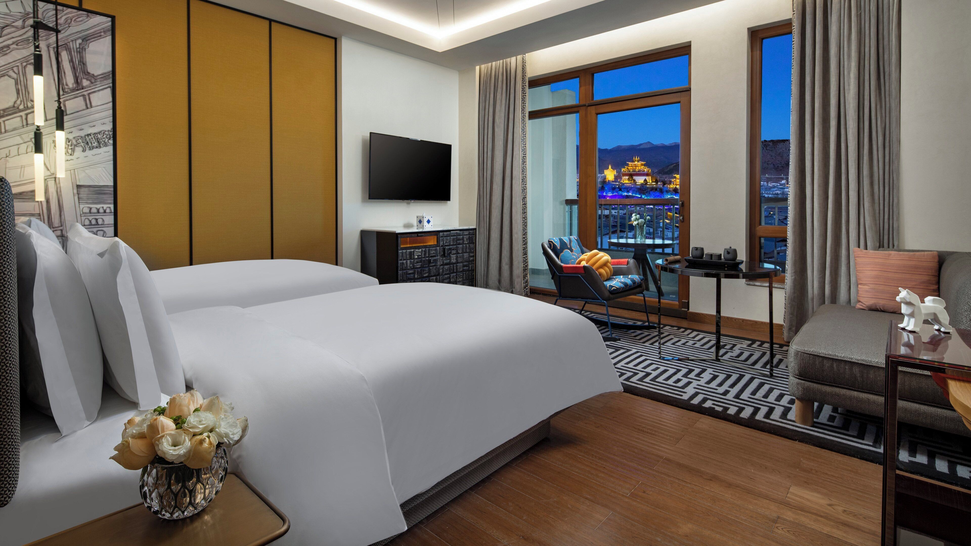 A hotel room with a large bed, a TV, a desk with a chair, and a view of a cityscape at night.