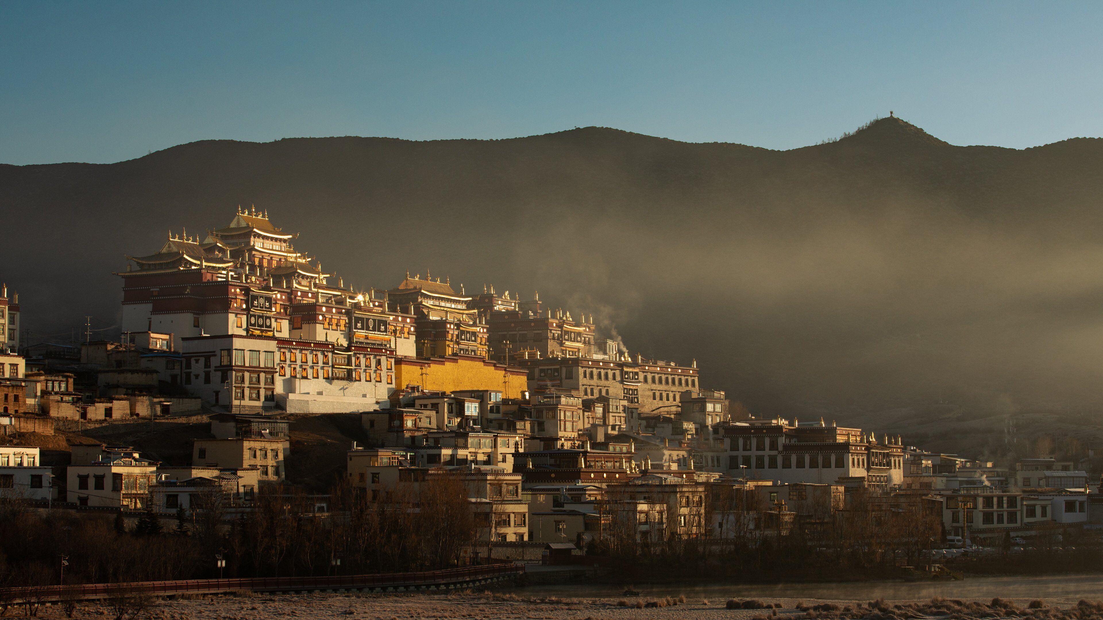 A traditional multi-story building with a complex roof structure, likely a monastery or cultural site, surrounded by a hilly landscape.