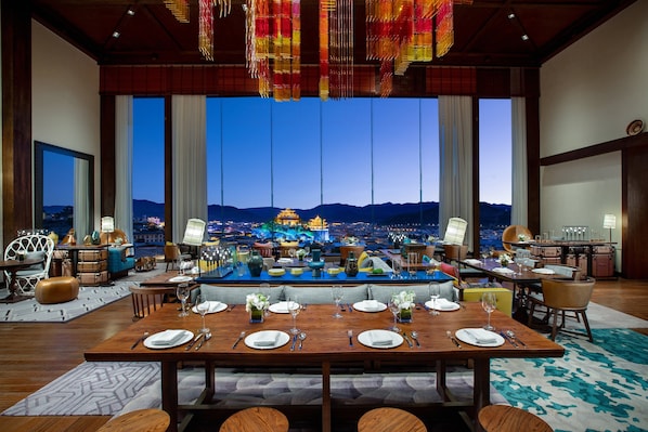 Comedor amplio con una mesa grande dispuesta para seis personas, zona de bar y vista a un paisaje urbano al anochecer.