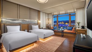 A hotel room with two beds, a sofa, a TV, and a view of a cityscape with illuminated buildings.