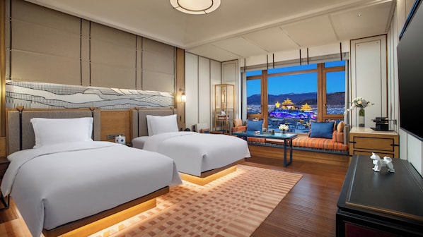 A hotel room with two beds, a sofa, a TV, and a view of a cityscape with illuminated buildings.