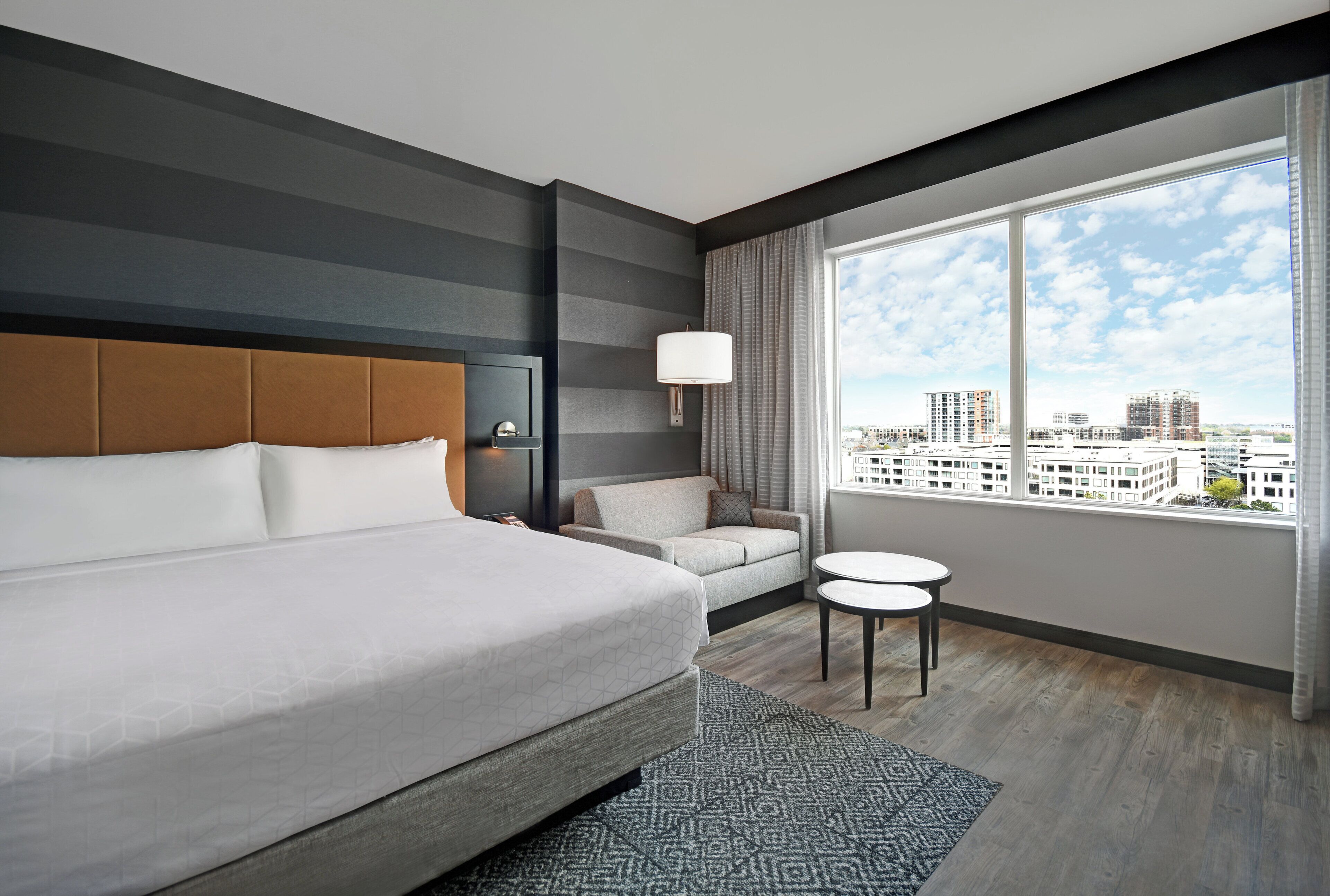 Quarto de hotel moderno com uma cama grande, um sofá, uma mesinha e vista para a paisagem urbana.