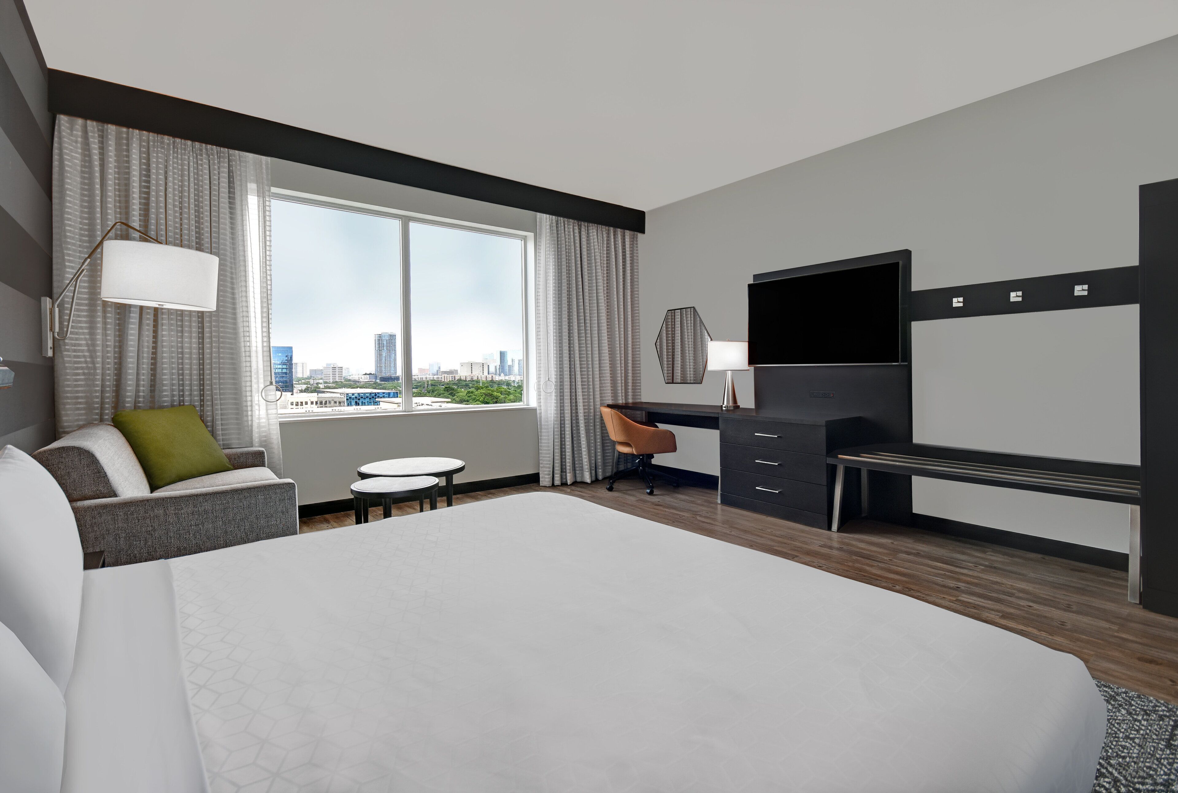 A modern hotel room with a large bed, a desk, a chair, a television, and a view of the cityscape.