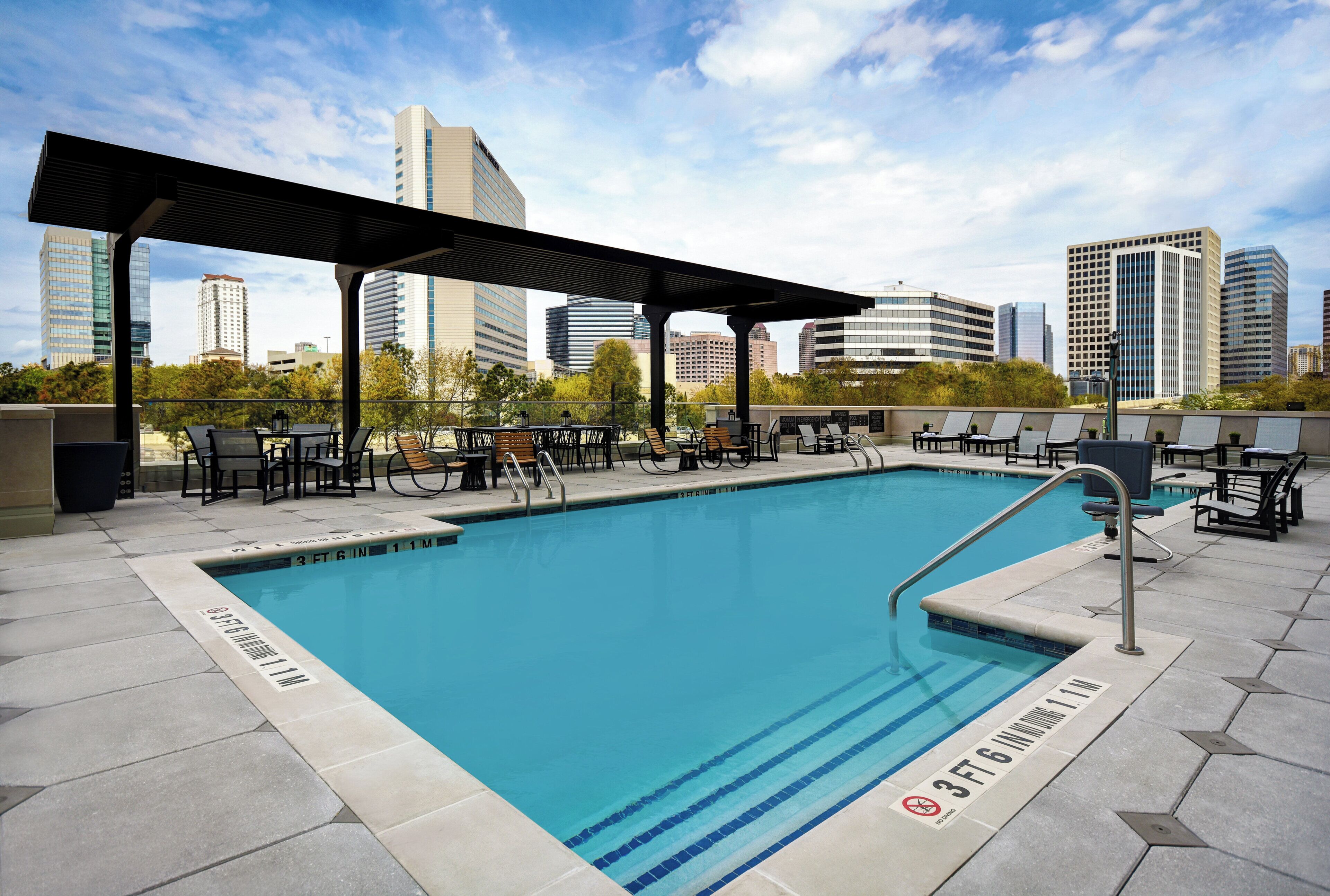 A rooftop pool with a city skyline in the background, surrounded by lounge chairs and tables.