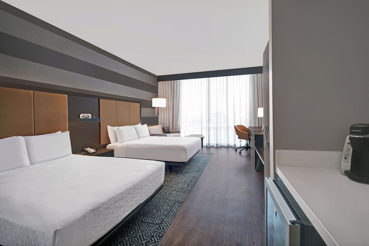 Ein Hotelzimmer mit zwei Betten, einem Schreibtisch, einem Stuhl, einer Kaffeemaschine und einem großen Fenster mit Vorhängen.