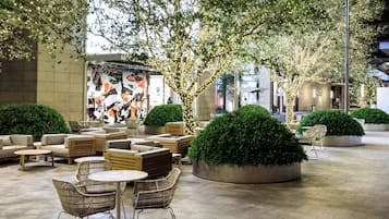 An outdoor seating area with round tables and wicker chairs, surrounded by lush greenery and trees.