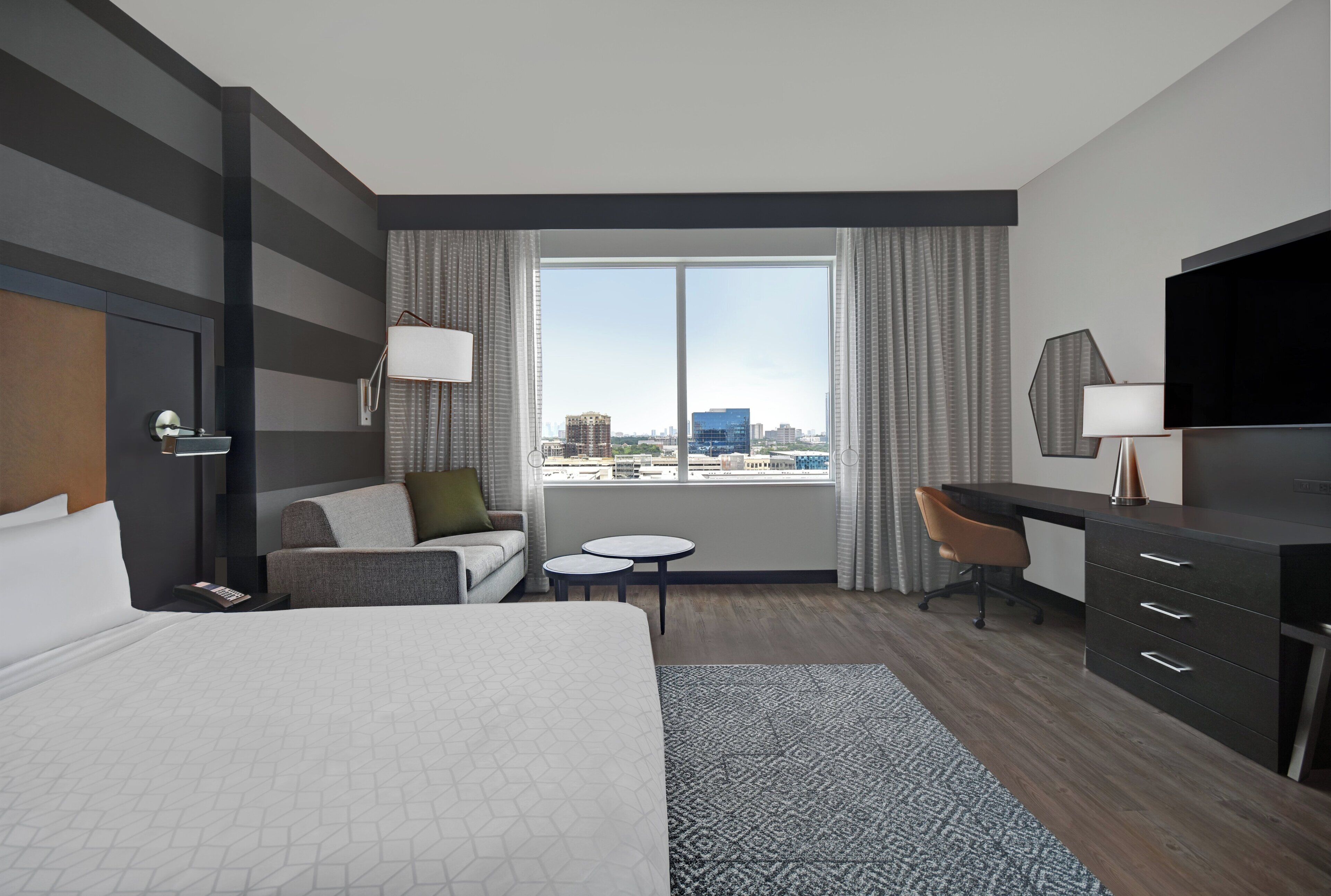 Ein Hotelzimmer mit Bett, Schreibtisch, Stuhl, kleinem Tisch und einem großen Fenster mit Blick auf die Stadt.