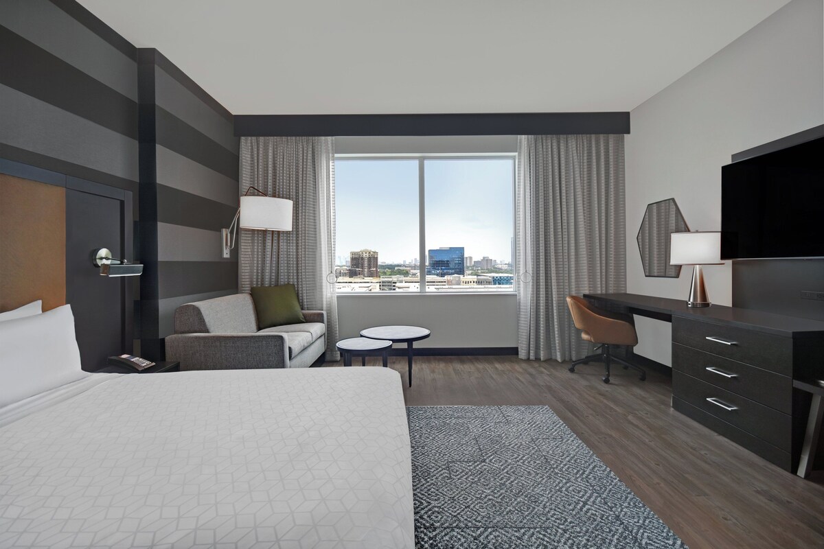 Ein Hotelzimmer mit Bett, Schreibtisch, Stuhl, kleinem Tisch und einem großen Fenster mit Blick auf die Stadt.