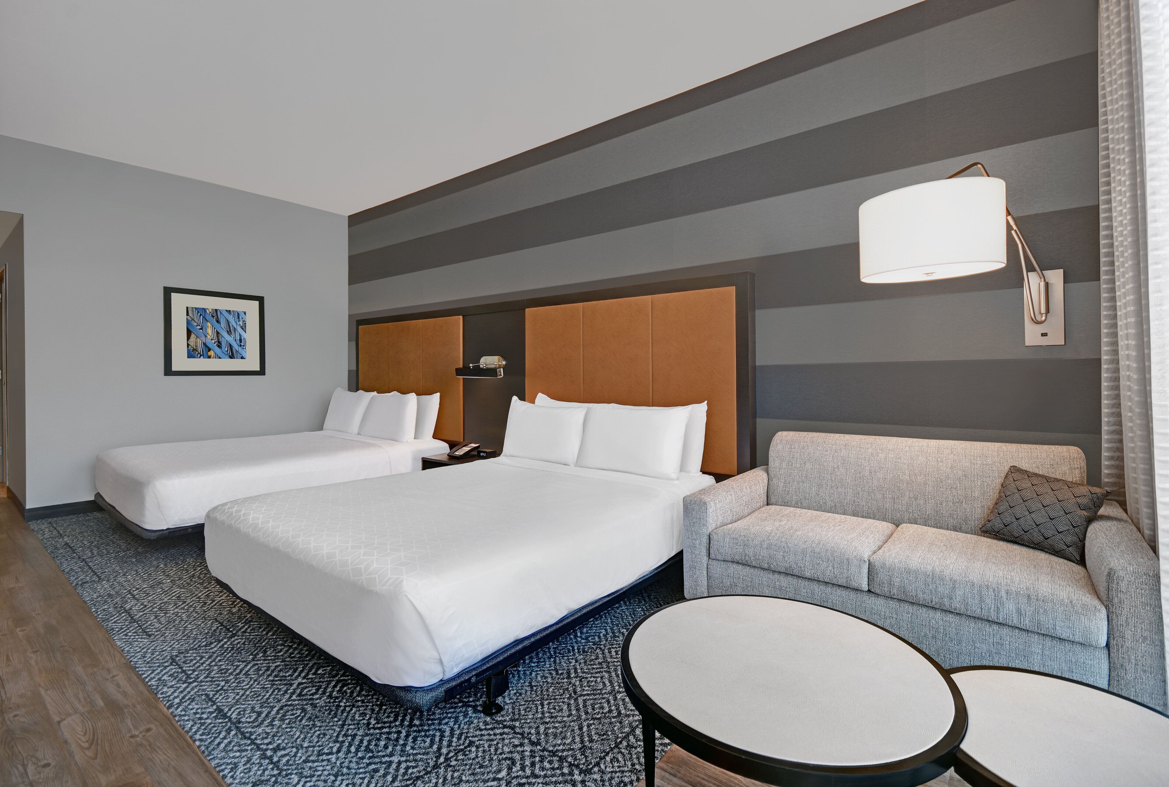 A hotel room with two beds, a sofa, a round table, and a wall-mounted lamp.