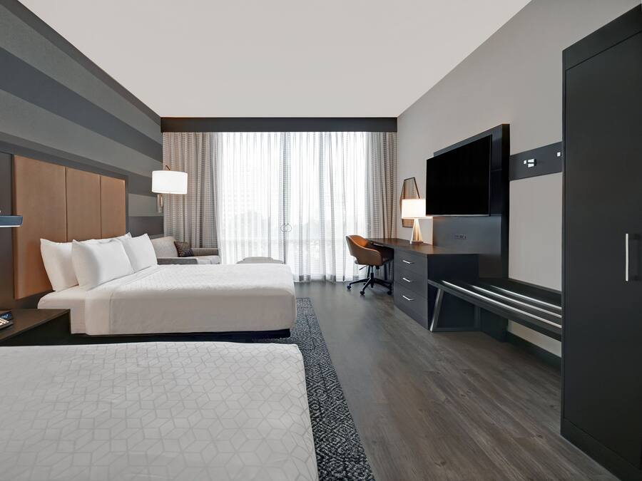 Habitación de hotel con dos camas, un escritorio, un televisor y un amplio ventanal con cortinas.