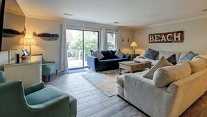 Un salon avec un canapé bleu, un canapé beige et un fauteuil vert. On y trouve une table basse et une lampe sur une table d’appoint. Une grande fenêtre avec des rideaux est visible, et un panneau sur le mur indique « PLAGE ».