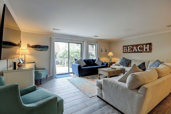 Una sala de estar con un sofá azul, un sofá beige y un sillón verde. Hay una mesa de centro y una lámpara sobre una mesita auxiliar. Se ve una ventana grande con cortinas, y un letrero en la pared dice "BEACH".