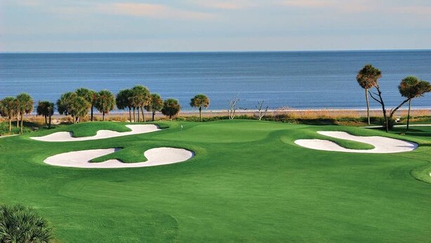 Un parcours de golf avec des bunkers de sable et des palmiers, offrant une vue sur l’océan.