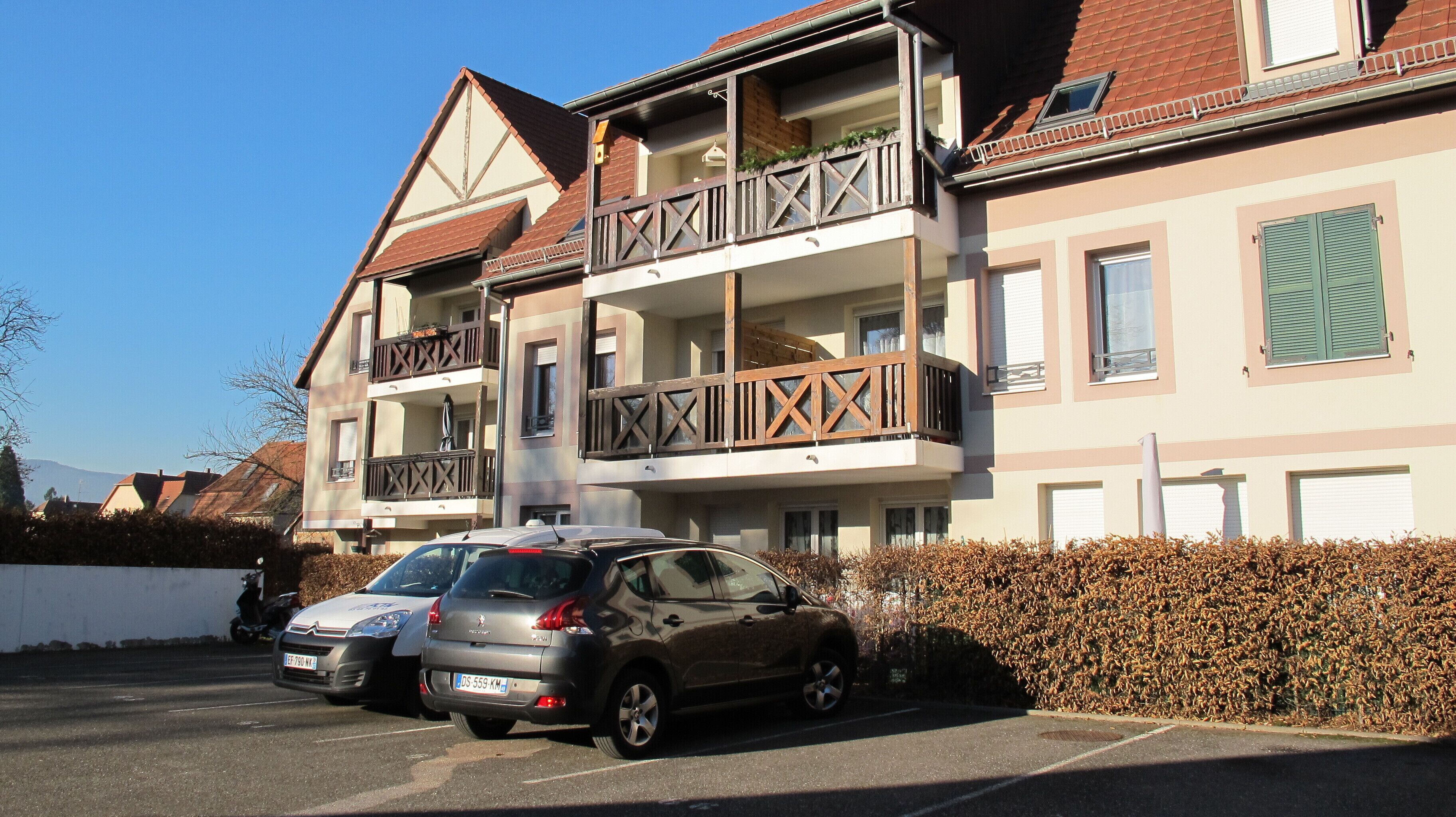 A multi-story residential building with balconies, a parking area, and a hedge.