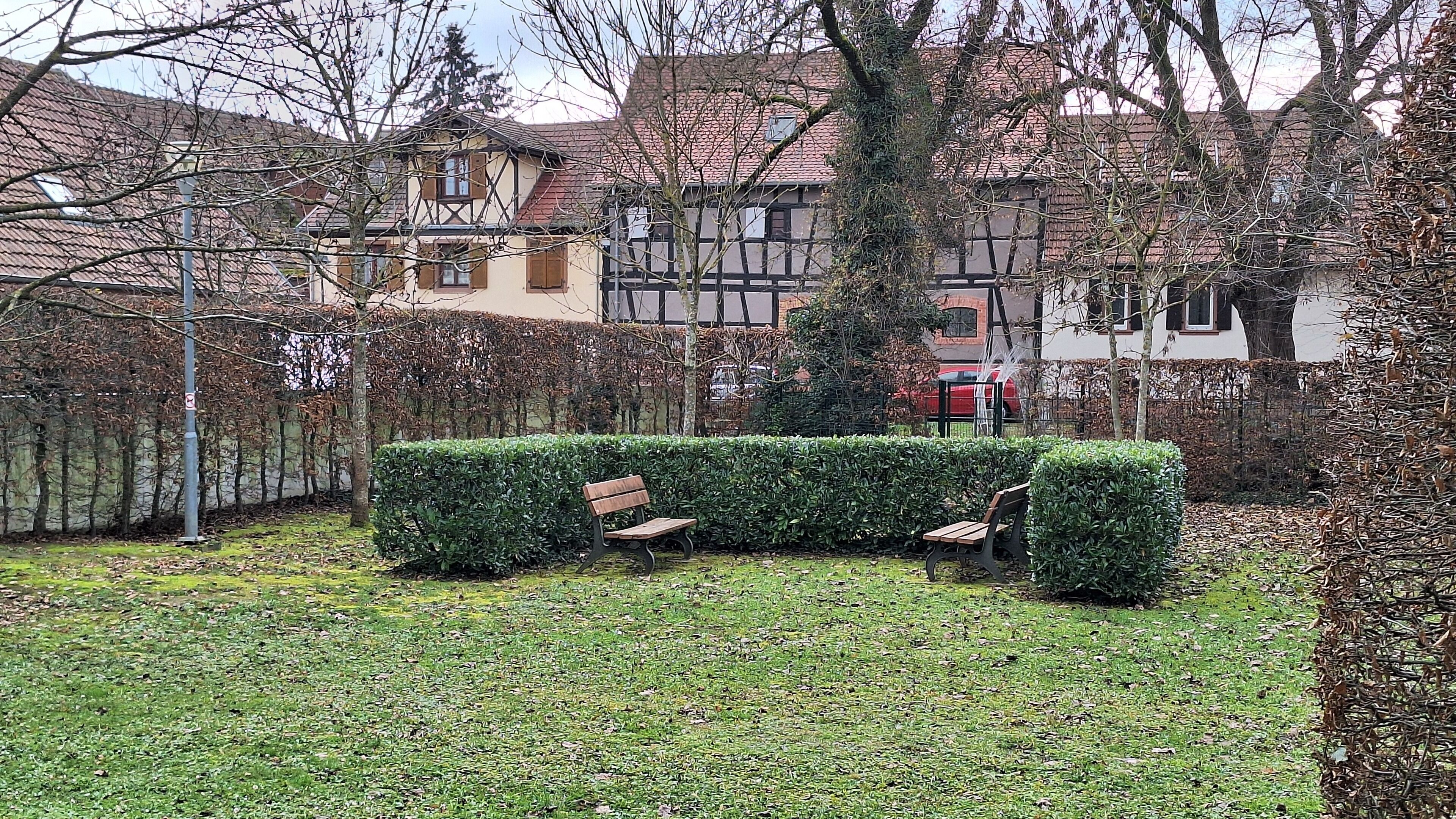 A courtyard with a bench, a hedge, and a red car parked in the background.