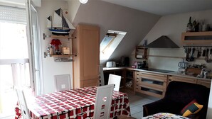 Et kompakt køkken med spiseplads, med et bord dækket af ternet dug, en stol og en seng med farverigt sengetæppe.