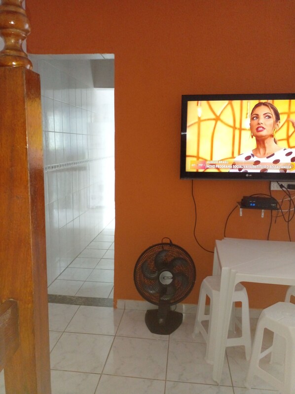 A room with a television mounted on an orange wall, a white table, and a black fan on the floor. - Casas em Condomínio Fechado ,com Tudo Perto ,comercio há 400 mts da Praia (SP)