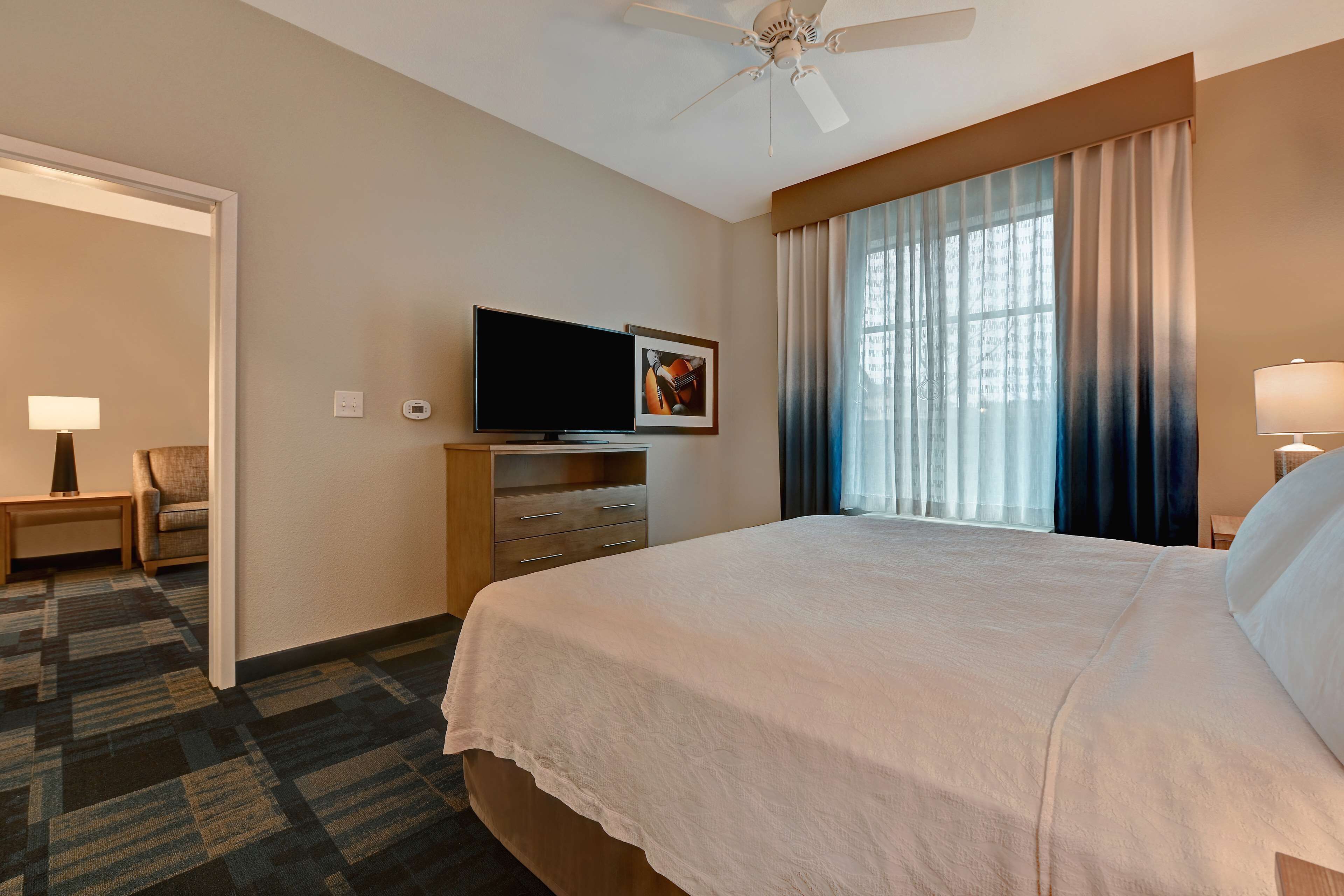 A hotel room with a large bed, a flat-screen TV, a desk with a lamp, and a window with curtains.