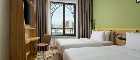 A hotel room with two beds, a desk, a chair, a TV, and a view of a cityscape through a large window.