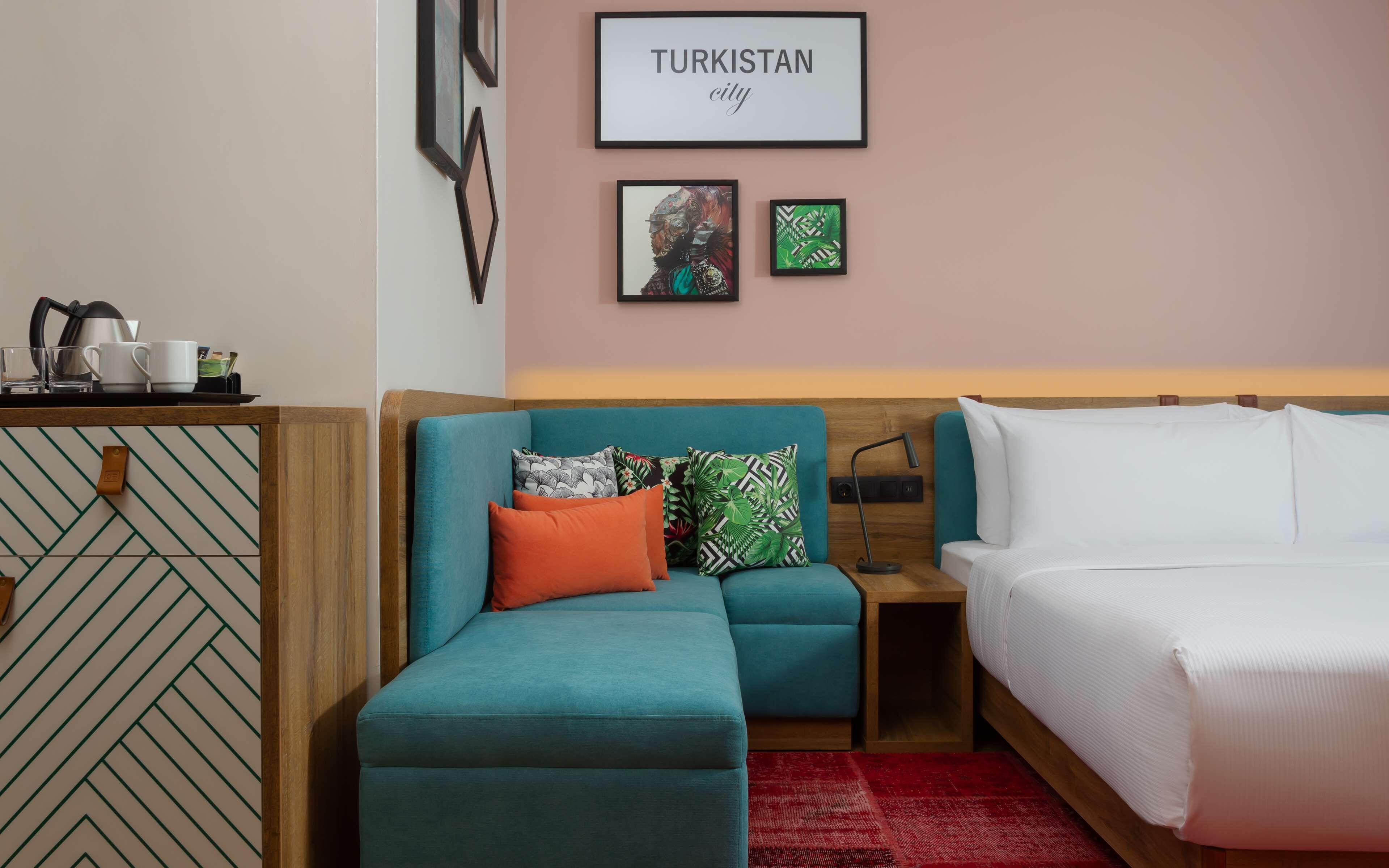 Habitación de hotel con un sofá color turquesa, una cama y una pared con cuadros, incluyendo uno con la etiqueta 'TURKISTAN'.