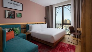 Une chambre d’hôtel avec un lit, un canapé, un bureau et une chaise. Deux tableaux encadrés sont accrochés au mur, l’un représentant un paysage urbain et l’autre, une plante verte.