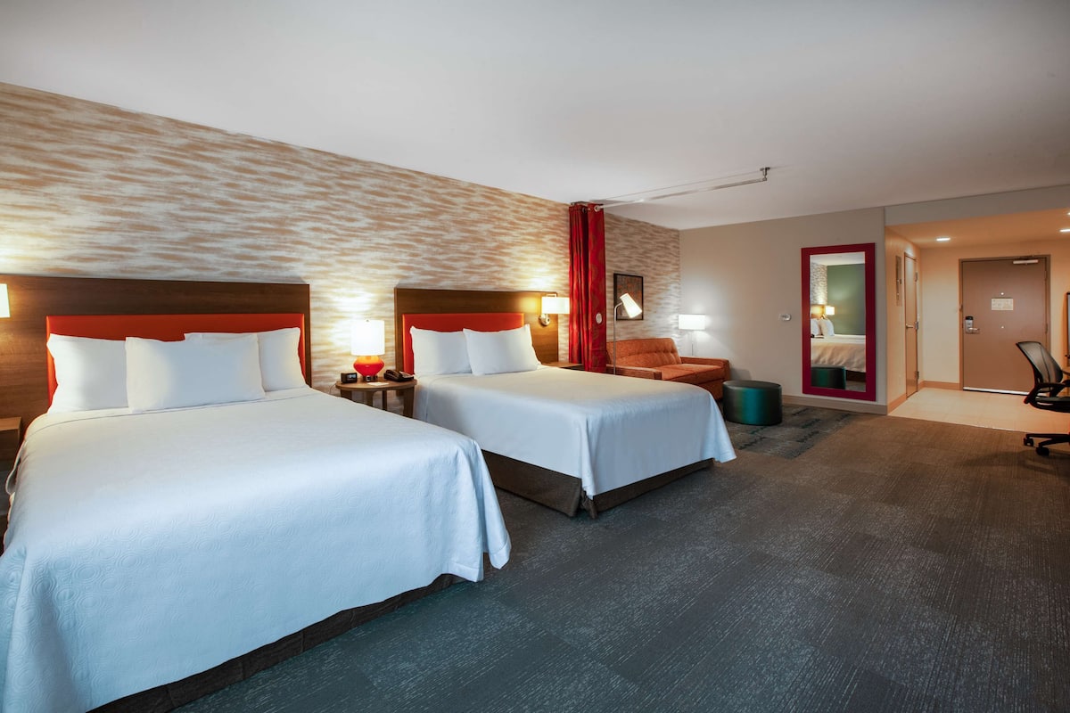 Habitación de hotel con dos camas, cabecera y cortina rojas, un escritorio con lámpara y una silla.