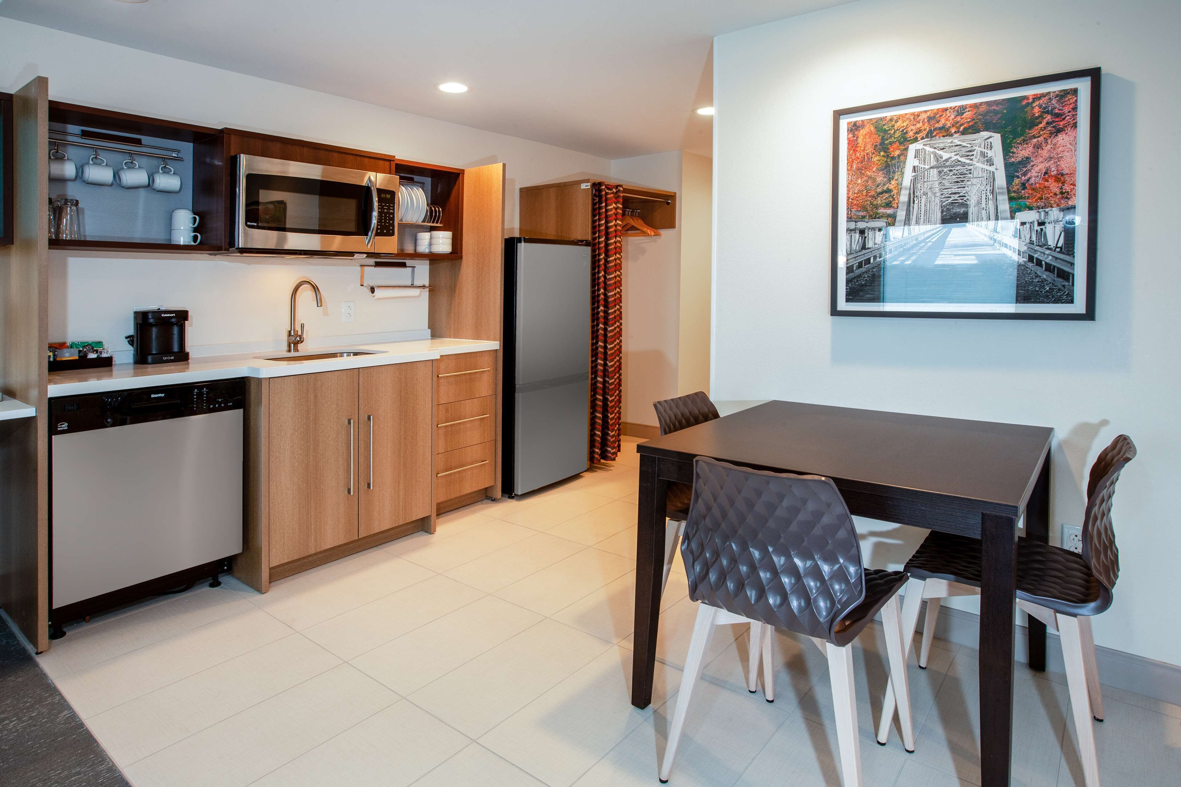 A modern kitchen with a dishwasher, microwave, and refrigerator. There is a dining table with chairs and a framed picture on the wall.