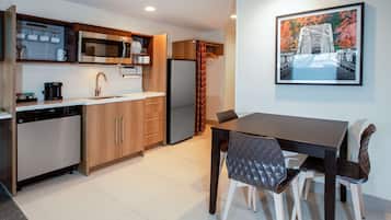 A modern kitchen with a dishwasher, microwave, and refrigerator. There is a dining table with chairs and a framed picture on the wall.