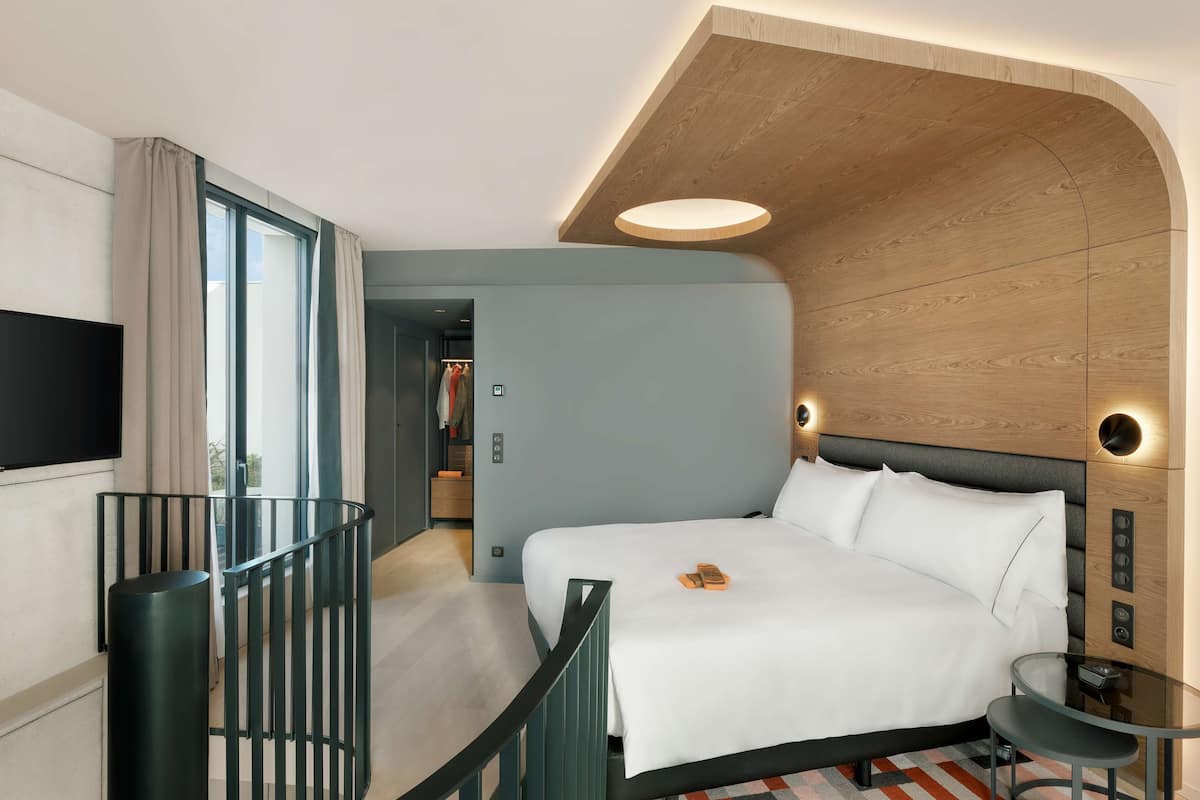 A modern hotel room with a large bed, a TV, a glass table, and a balcony with a railing.