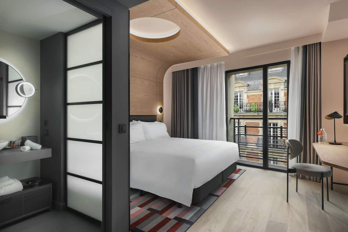 A modern hotel room with a large bed, a desk, a chair, a mirror, and a view of the city through large windows.