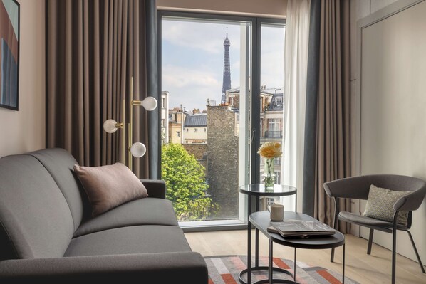 Ein modernes Wohnzimmer mit einer Couch, einem kleinen Tisch und einem Sessel. Ein großes Fenster bietet einen Blick auf die Stadtlandschaft, darunter ein hoher Turm.
